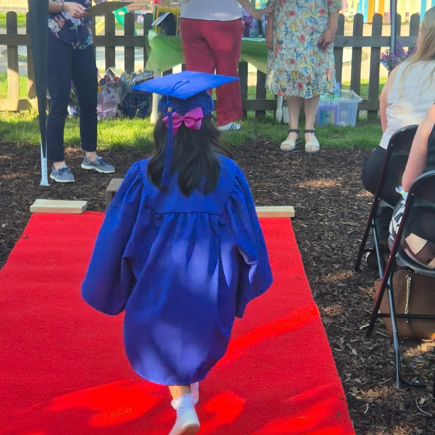 A preschool graduate in a blue cap and gown walking along a red carpet, seen from behind